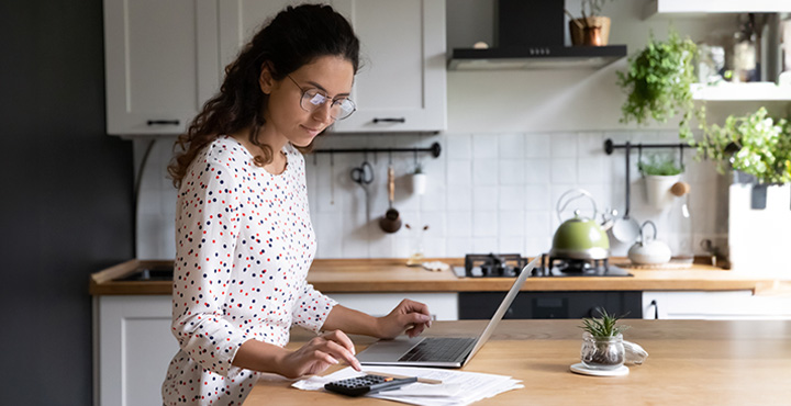 Person in their kitchen is on their laptop with a calculator nearby looks where they can grow financially.