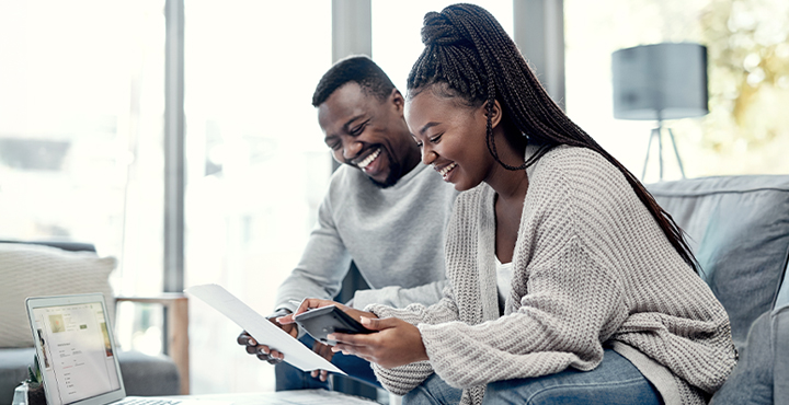 Couple sitting on a couch smiling looking at a smart phone and laptop